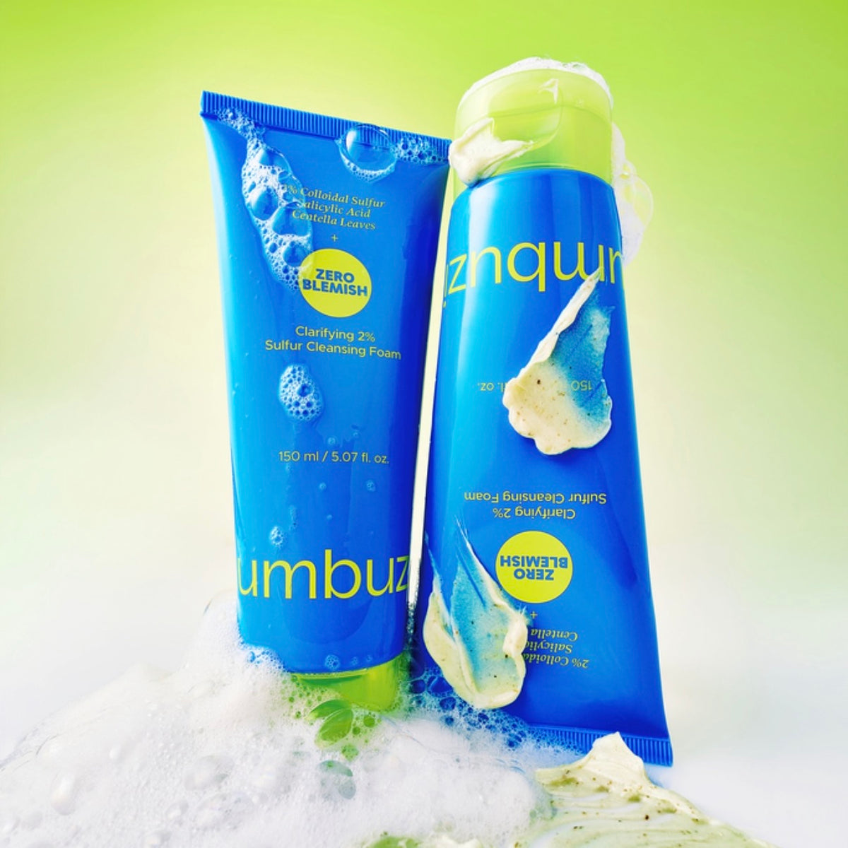 Two blue tubes of numbuzin Zero Blemish Clarifying 2% Sulfur Cleansing Foam, 150 ml, leaning together, with white foam below and dabs of cream cleanser on the tubes, against a yellow-green background.