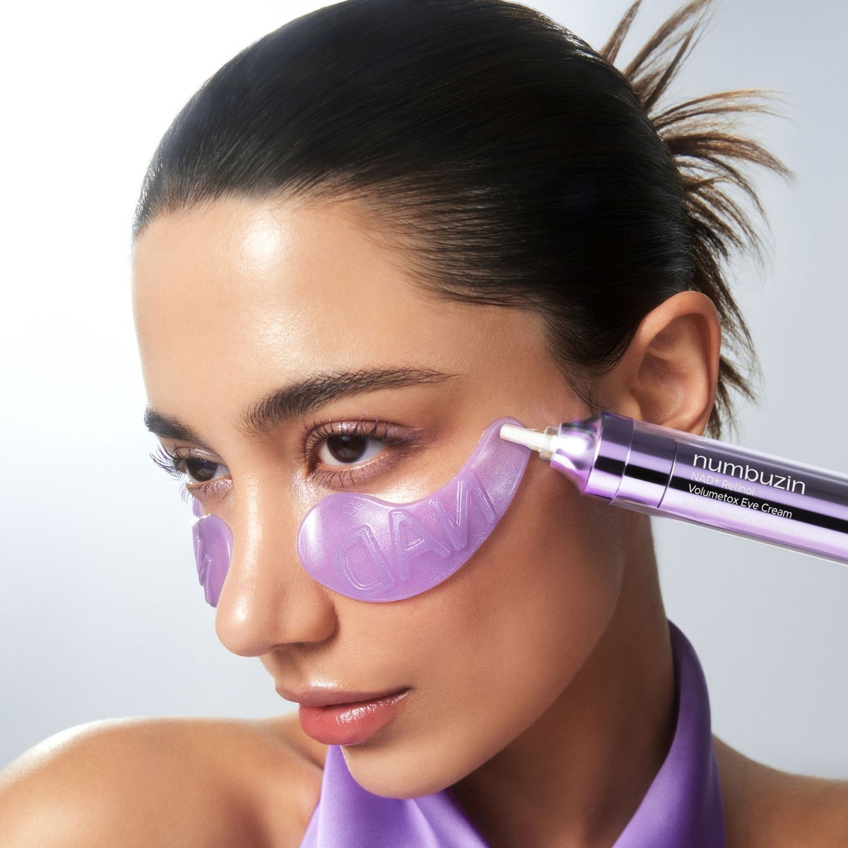 Close-up of a face with smooth, dewy skin wearing purple NAD under-eye patches while a numbuzin NAD+ Retinol Volumetox Eye Cream applicator touches the cheek beside the patch, against a soft gray background.